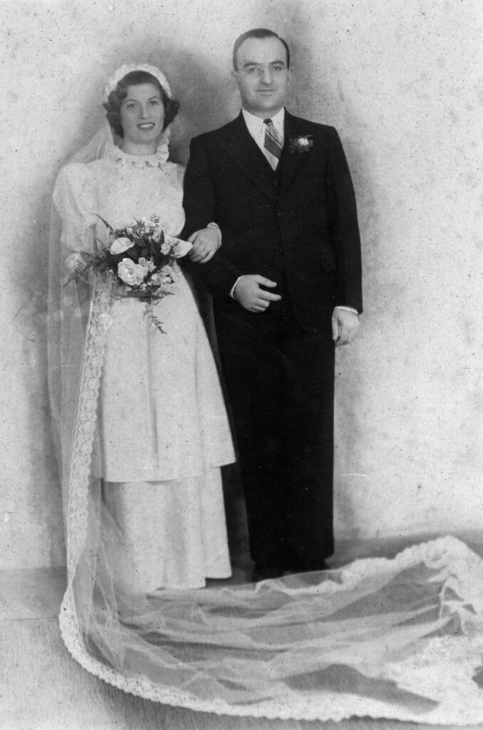 Black and white photo of bride in lacey dress with ruff and puff sleeves with very long lacey veil headdress and bouquet arm-in-arm with groom in suit