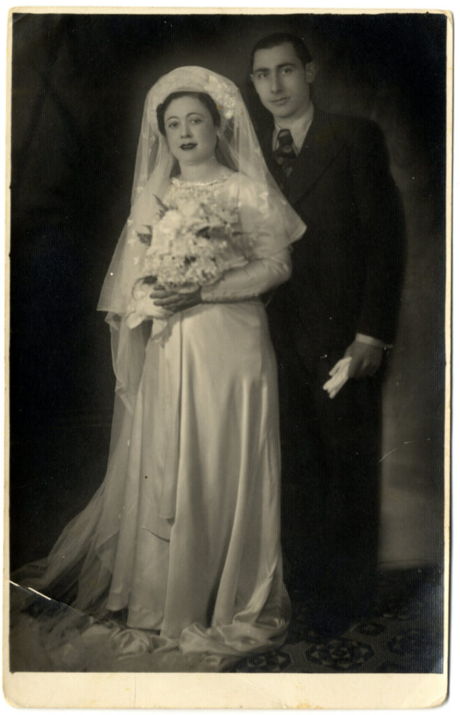 Black and white portrait of bride in shiny long dress with long sleeves holding grand bouquet wearing large hat with veil running down to floor; and tall groom wearing suit, holding handkerchief