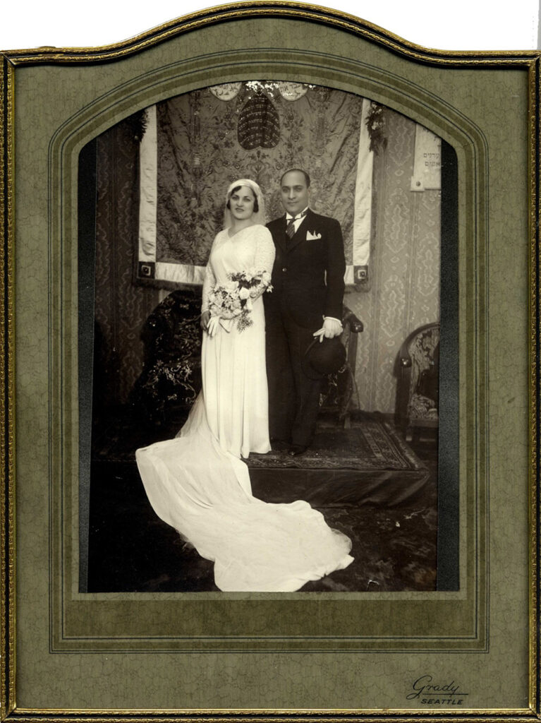 Black and white portrait of bride in elegant white dress with flowing train and groom in suit standing on platform with a tapestry and lavish Ottoman-style chairs and carpet around