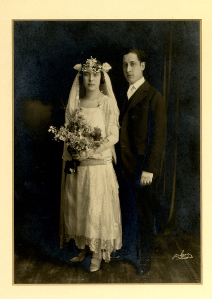 Black and white photo of bride in lacy dress and long gloves, veil headband and bouquet, with groom in suit and gloves