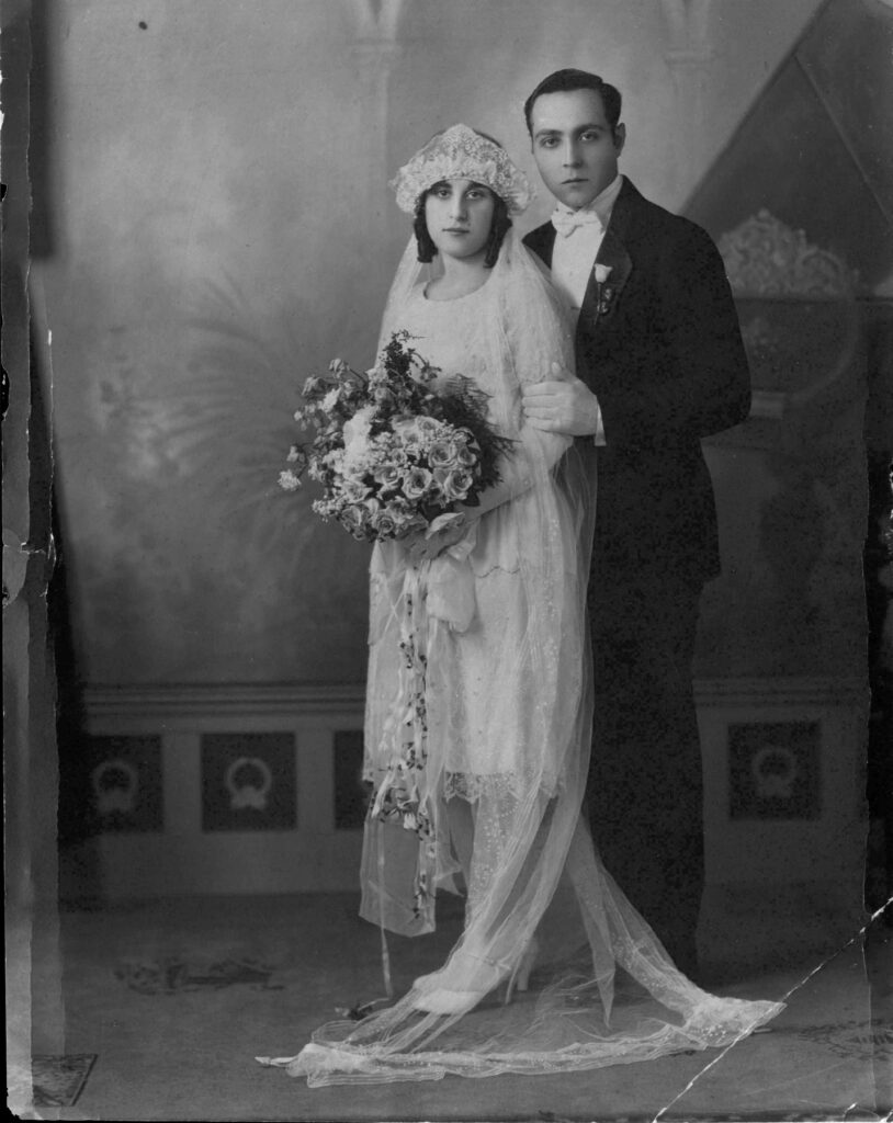 Black and white portait of bride in short lacey dress wearing long veil headdress, groom in suit, in ornate space with marble floor