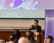 A presenter at the podium facing the front of a crowd. Behind is a projector screen with the words "Rafael Neis, University of Michigan"