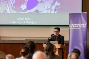 A presenter at the podium facing the front of a crowd. Behind is a projector screen with the words "Rafael Neis, University of Michigan"