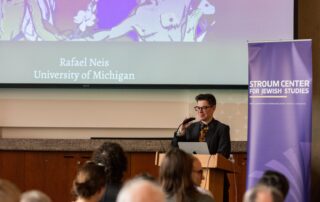 A presenter at the podium facing the front of a crowd. Behind is a projector screen with the words "Rafael Neis, University of Michigan"