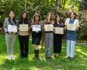 Six students smile for a group photo in a leafy outdoor setting while displaying certificates recognizing their Student Research Symposium achievements.