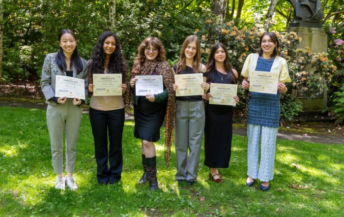 Six students smile for a group photo in a leafy outdoor setting while displaying certificates recognizing their Student Research Symposium achievements.