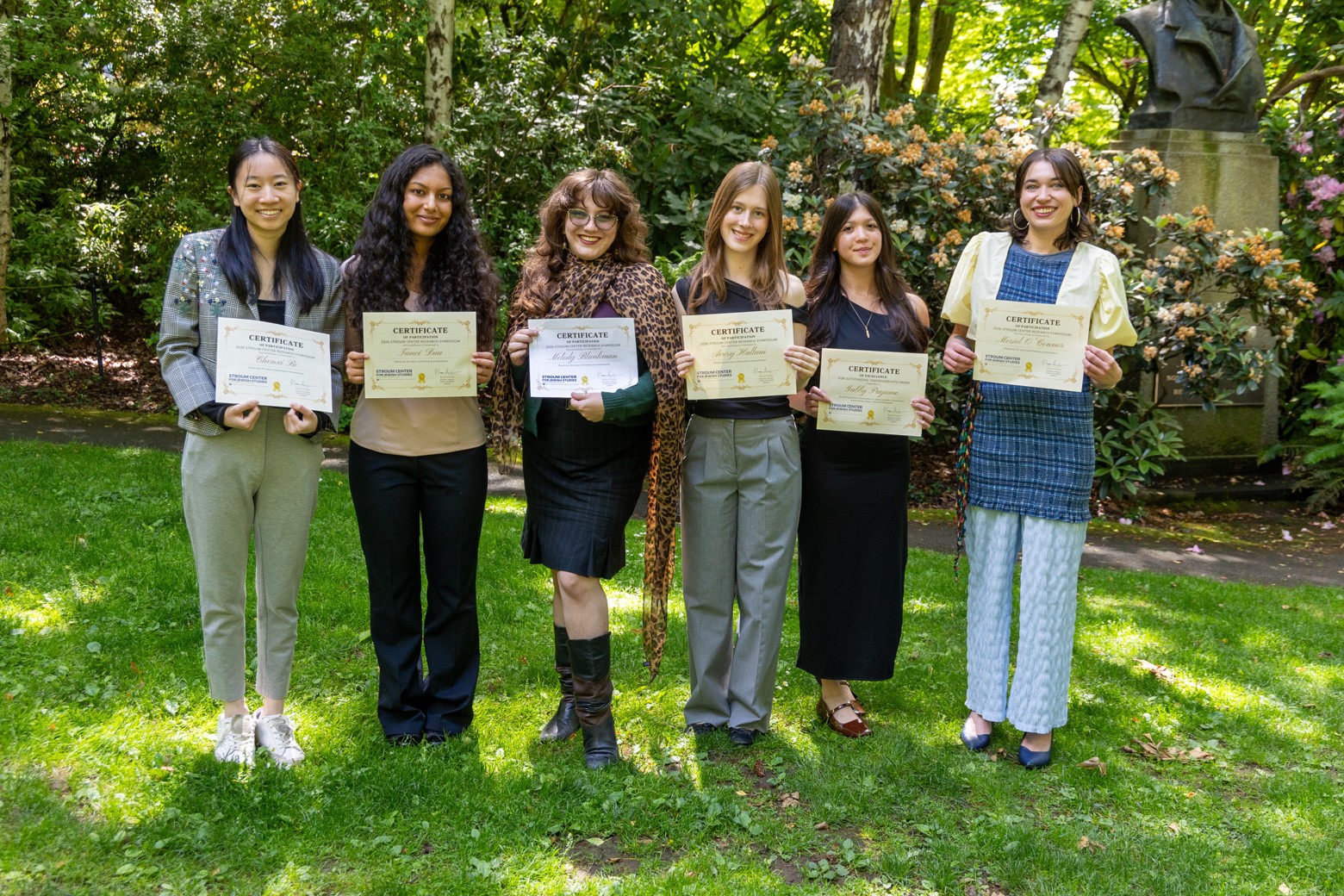 Six students smile for a group photo in a leafy outdoor setting while displaying certificates recognizing their Student Research Symposium achievements.