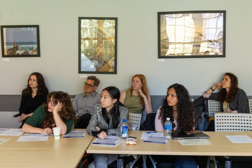 A group of symposium attendees sits with handouts, notebooks, and water bottles while looking toward a speaker off camera.