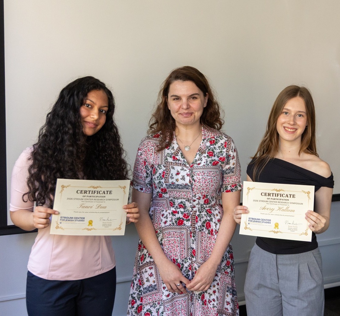 Three people pose indoors in front of a projection screen, with two holding certificates from a research symposium.