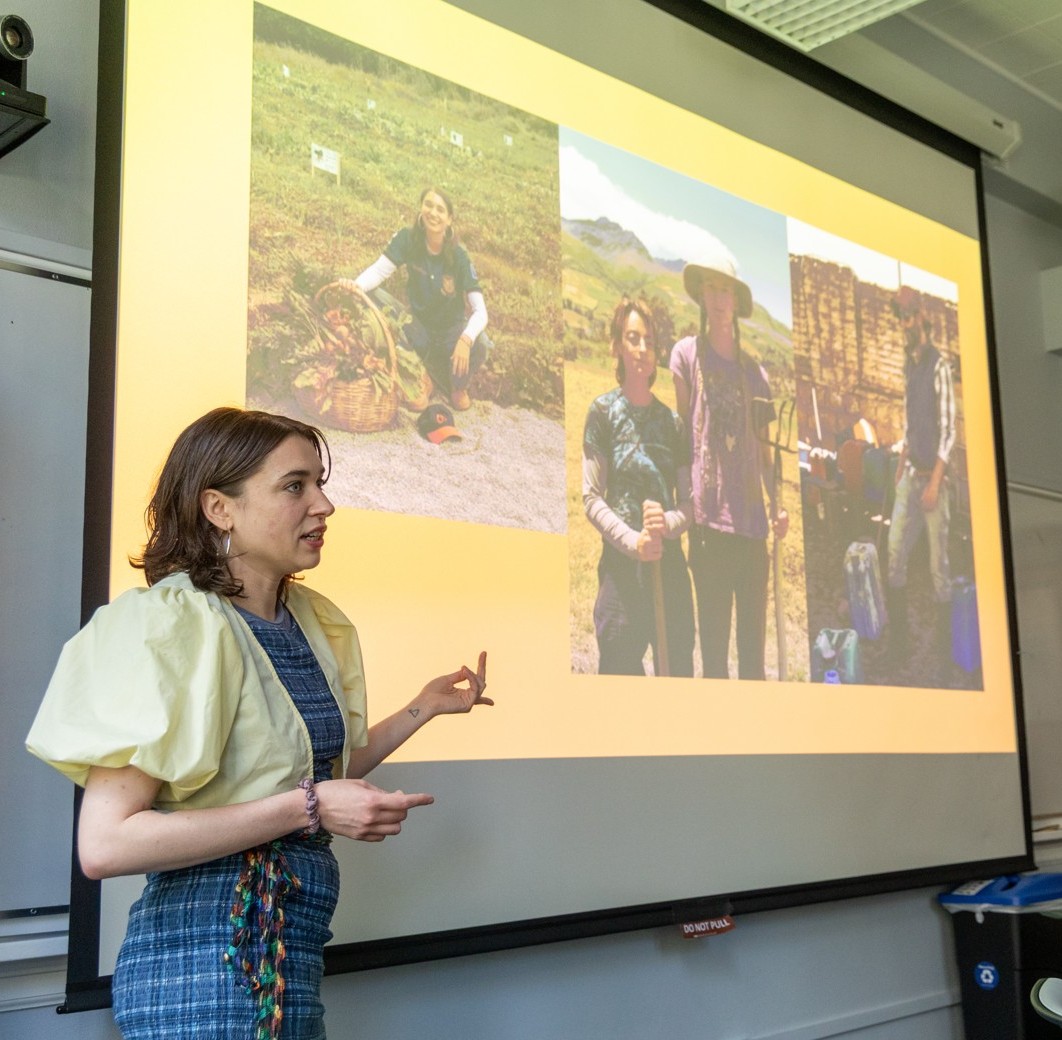 A presenter gestures toward a slideshow with fieldwork images during a research symposium presentation in a classroom.