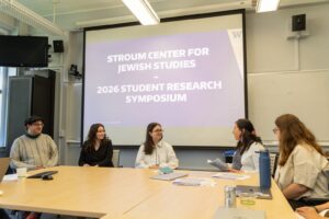 A group of symposium participants sits around a meeting table with papers, laptops, and drinks, while a presentation slide for the Stroum Center for Jewish Studies is projected behind them.