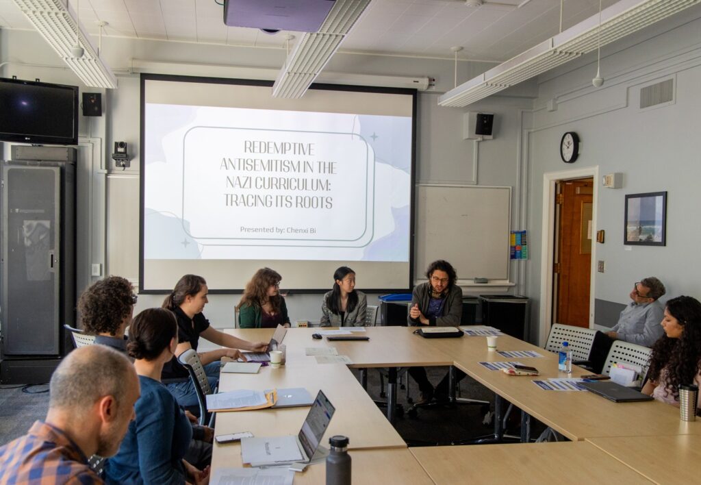 People sit around a conference table during a student research symposium presentation, with a projected slide titled “Redemptive Antisemitism in the Nazi Curriculum: Tracing Its Roots.”