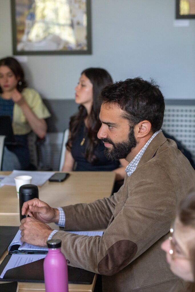 A man in a brown jacket sits at a conference table listening during a classroom discussion, with other participants seated nearby.