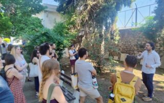 Students gather in a leafy outdoor space while listening to a speaker standing near a stone wall and trees.