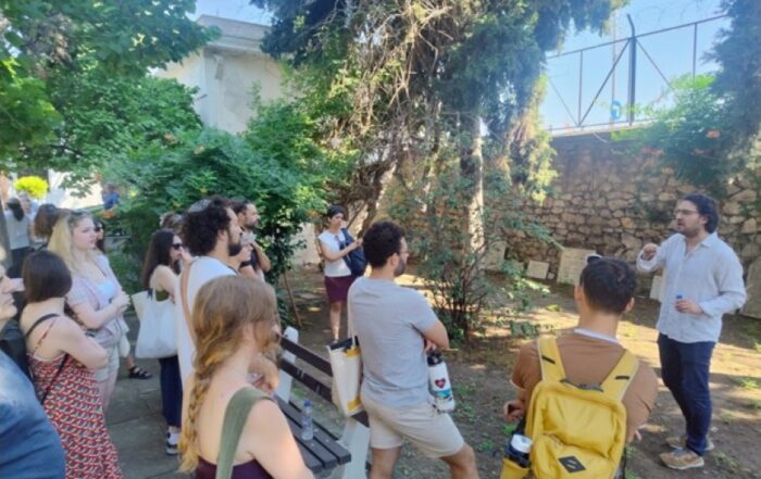 Students gather in a leafy outdoor space while listening to a speaker standing near a stone wall and trees.