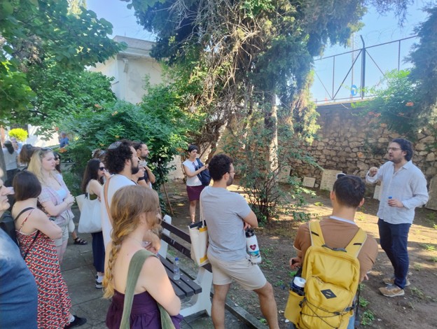 Students gather in a leafy outdoor space while listening to a speaker standing near a stone wall and trees.