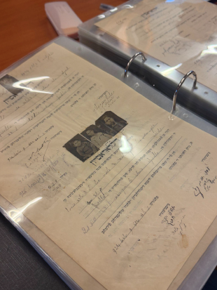 Archival papers with handwritten annotations and a small photograph are stored in clear protective sleeves inside a ring binder on a table.