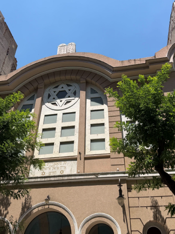 The exterior facade of a synagogue with a large Star of David window, arched architectural details, Hebrew inscription, and trees in front.