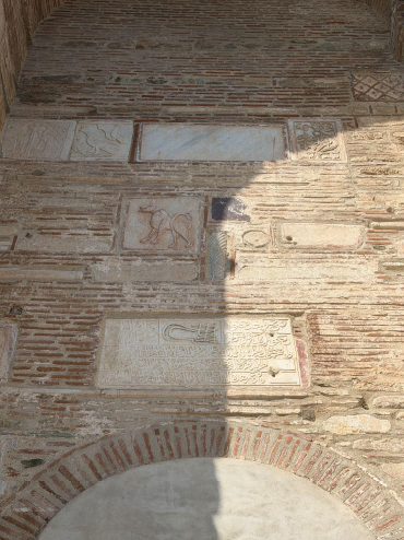 Close-up of an old masonry wall with marble fragments set into the brickwork, including carved animal figures, inscriptions, and ornamental designs.