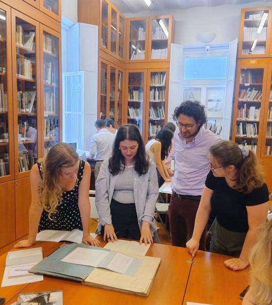 Four people stand around archival materials on a table while others work in the background, surrounded by tall glass-front bookcases.