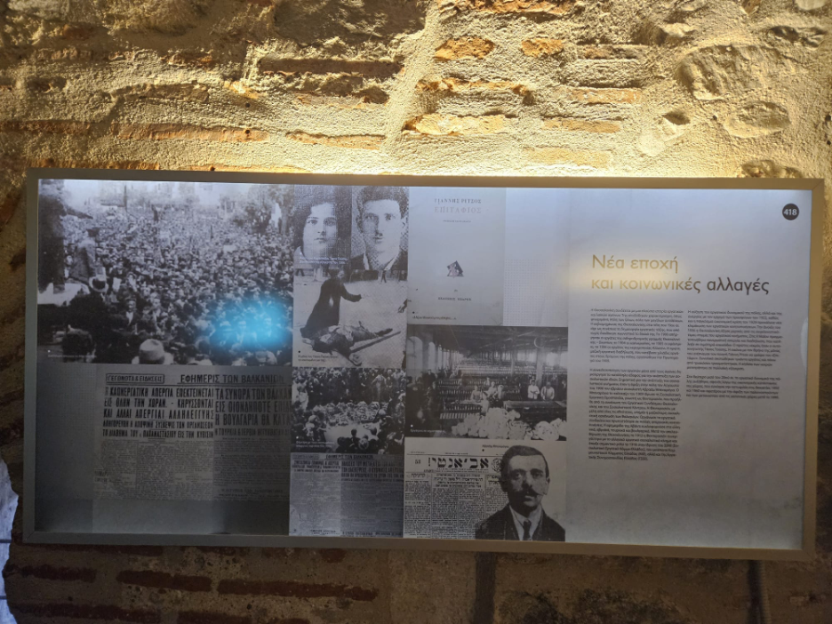 A historical museum exhibit presents a collage of archival photos and documents beside a section of Greek text, set against a rough stone wall.