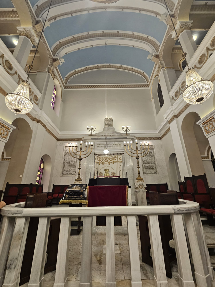 Interior view of a synagogue featuring a central bimah, tall menorahs, decorative chandeliers, marble walls, and Hebrew text panels.