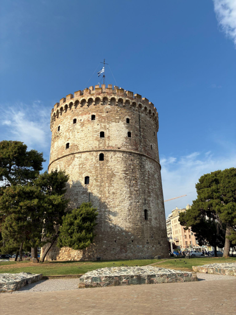 The White Tower of Thessaloniki rises above a park-like area with trees, stone ruins, and a clear blue sky.