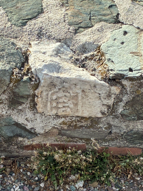 An old masonry wall contains an embedded rectangular stone piece with faint carved markings, surrounded by cracked mortar and small plants.