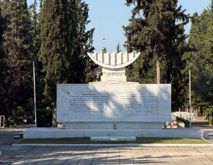 A large stone memorial with a menorah sculpture stands in a tree-lined plaza, with engraved text panels on the front.