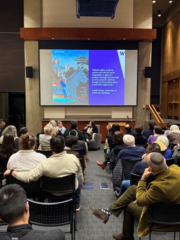 An audience watches two seated speakers in conversation beneath a projected slide at a book launch event.