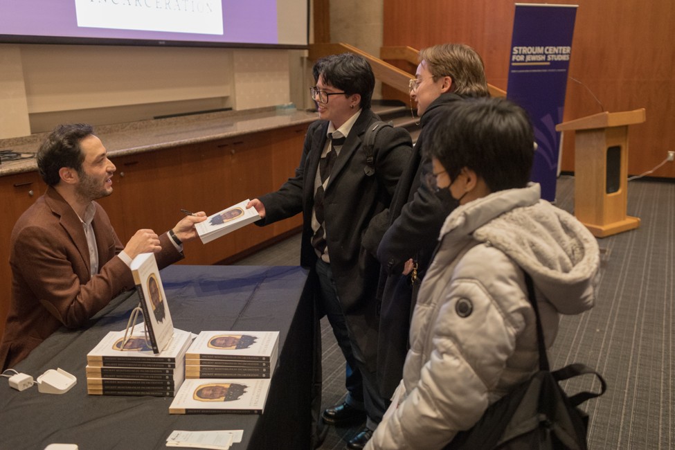 Attendees speak with a seated author at a table stacked with books during a book signing.