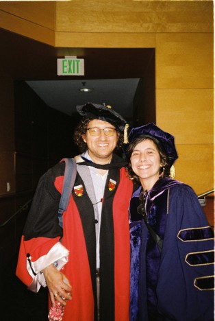 Two smiling graduates in academic regalia pose together indoors